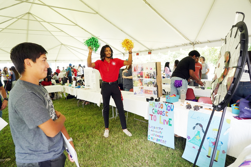 A young African American man in a red polo shirt and black pants waves green and yellow pom-poms under a tent with many tables set up beneath it and students milling around