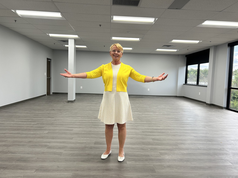 A blond, short-haired woman wearing a cream dress and yellow sweater stands with arms spread wide in the middle of a brand-new classroom